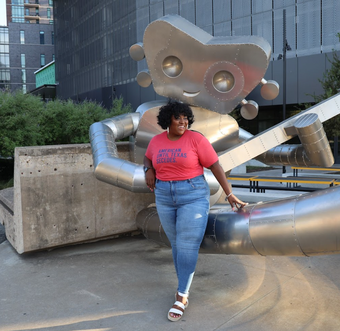 Person wearing an 'American until Texas Secedes' tshirt and blue jeans standing next to a large metallic Robot sculpture in Deep Ellum.