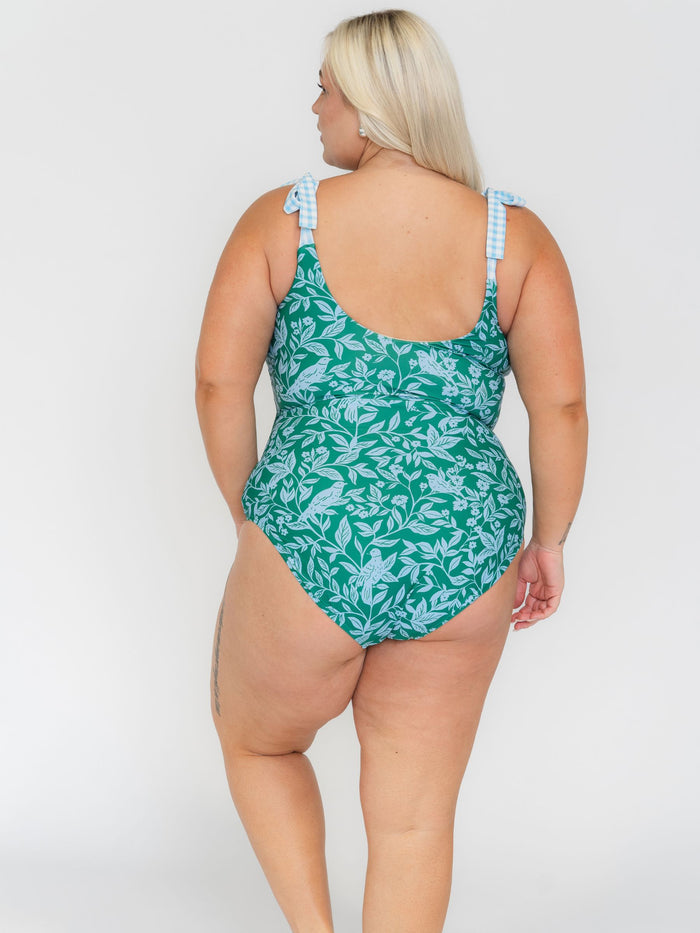 Woman wearing a white leaf-patterned one-piece swimsuit with blue and white checkered straps on a white background