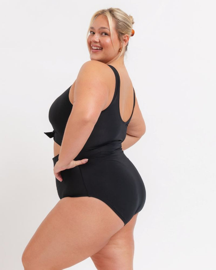 side view of a woman wearing a black knotted one piece with a white background