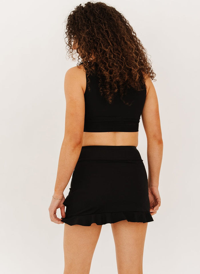 woman wearing a black high-waist ruffle swim skirt with a black knotted crop swim top back angle