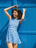 Woman in a blue gingham dress standing against a blue wooden wall.