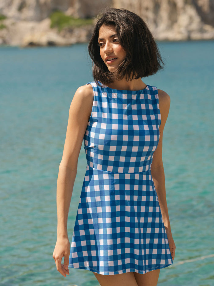 Woman wearing a blue gingham swim dress standing by a body of water.