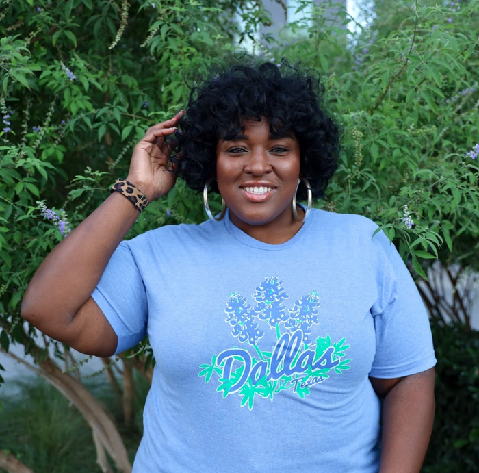 Photo of a woman wearing the Bullzerk Bluebonnets tshirt standing in front of greenery and smiling.