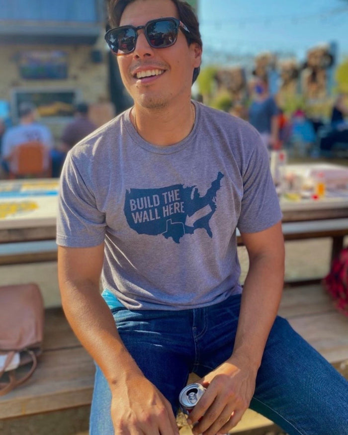 Man wearing a Bullzerk "Build the Wall Here" tshirt and sunglasses, sitting at a wooden picnic table and holding a can of beer.