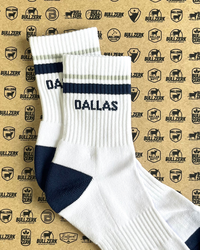 A pair of white crew socks with blue and red stripes and 'DALLAS' text laying on a patterned background.