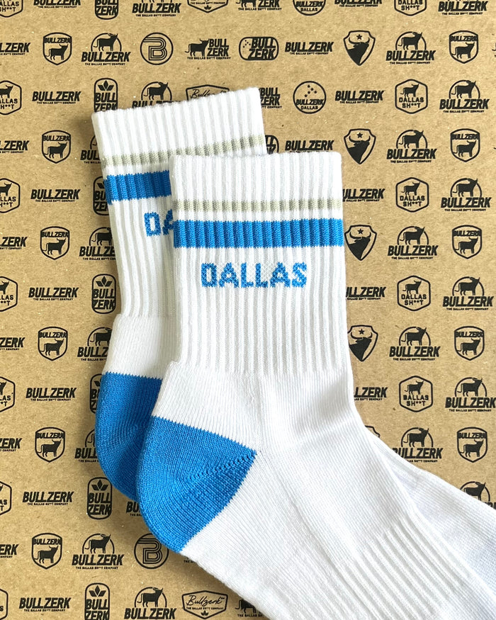 A pair of white crew socks with blue and gray stripes and 'DALLAS' text laying on a patterned background.