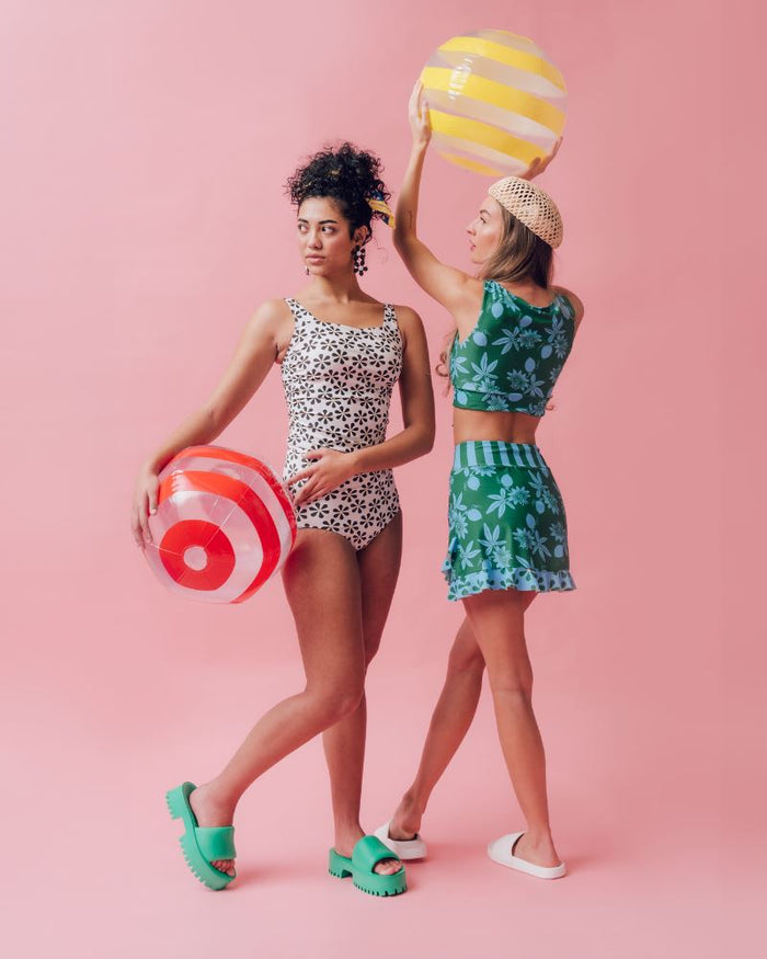 Photo of two women wearing a black and white floral square neck swim top and a black and white floral swim bottom and a green and blue floral swim crop top and a green and blue swim skirt bottom