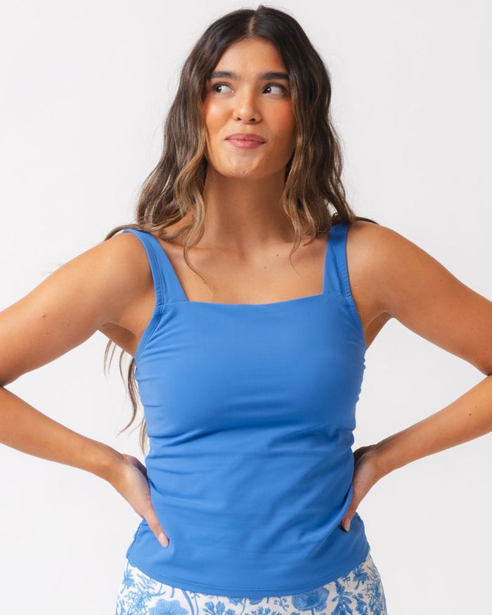 A woman modeling a bright blue tank top with a square neckline and wide shoulder straps, paired with blue-and-white patterned bottoms, against a clean white studio background.