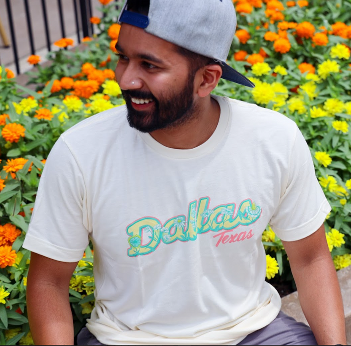 Photo of a man wearing the Bullzerk Dallas Flowers tshirt sitting next to a group of flowers and smiling.