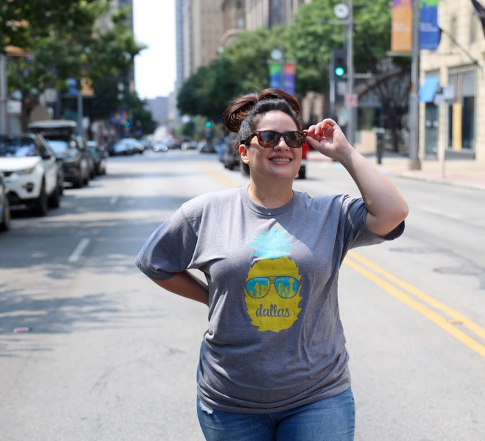 Photo of a woman wearing sunglasses and the Bullzerk Dallas Pineapple tshirt standing in the street outside.