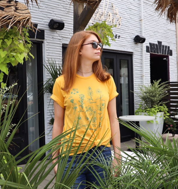 Woman standing near bushes wearing sunglasses and a Bullzerk Flowers of Texas tshirt.