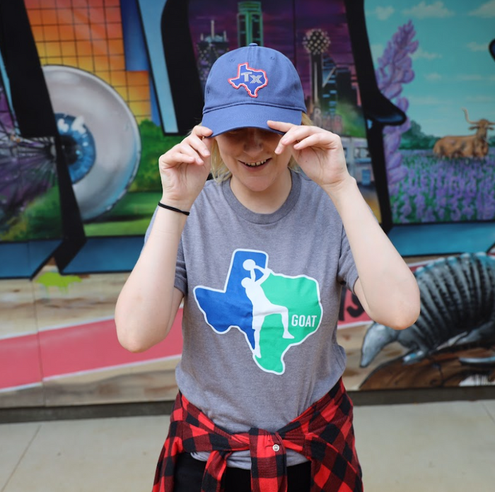 Woman wearing a Bullzerk GOAT tshirt and a Texas Silhouette Baseball hat, standing in front of an art mural.