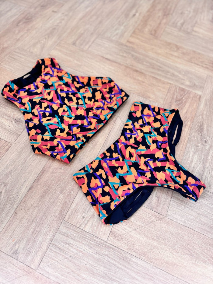 This image features the "Hitchhiker Bottoms- NOW & THEN" by Strange, modeled in a daring and playful spirit. The high-waisted bikini bottoms are showcased in a vibrant cantaloupe color, designed to reverse, as indicated, to a medina pattern.