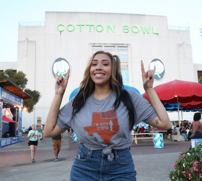 Woman posing in front of the Cotton Bowl stadium wearing a Bullzerk I'm With Stupid tshirt and making the Hook 'em Horns sign with her hands.