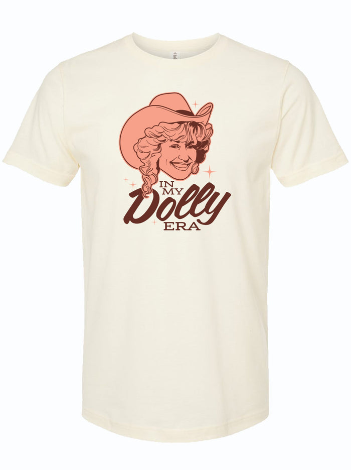 A cream shirt with a light orange and maroon illustration of Dolly Parton wearing a cowboy hat that reads "IN MY DOLLY ERA."
