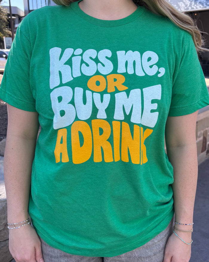 Person wearing a green tshirt with graphic design of text reading "kiss me, or buy me a drink" in yellow and white bubble letters.
