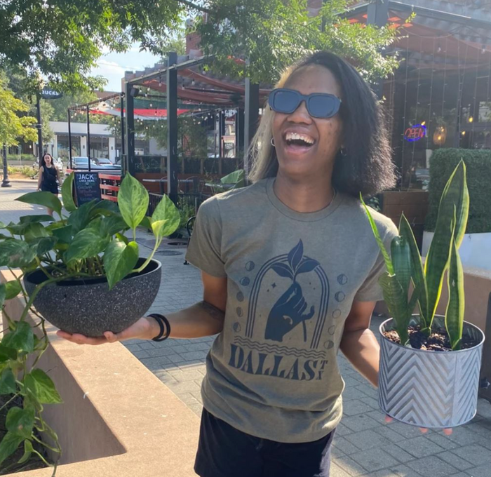 Woman standing outside, wearing a Bullzerk Mystic Dallas tshirt and holding two plants.