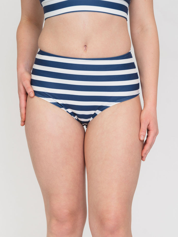 A model wearing a high-waisted bikini bottom with navy blue and white horizontal stripes, shown from the waist to the upper thighs against a light gray studio background.