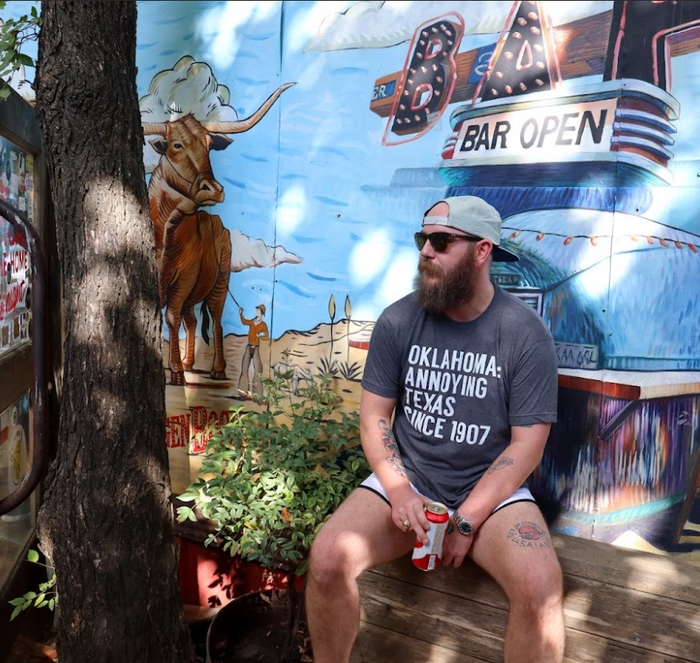 Man sitting on a bench in front of a colorful mural, wearing a Bullzerk Oklahoma Annoying tshirt.