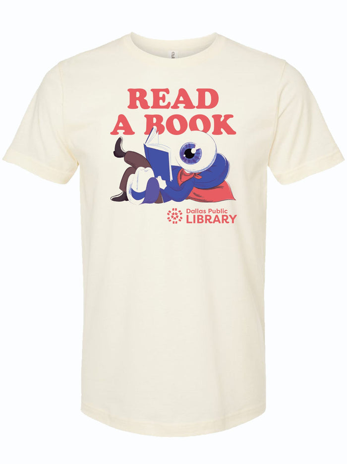 Cream tshirt with graphic design of a character with an eyeball as a head laying down reading a book. It is undereath red text that says "read a book" and above a logo for the Dallas Public Library.