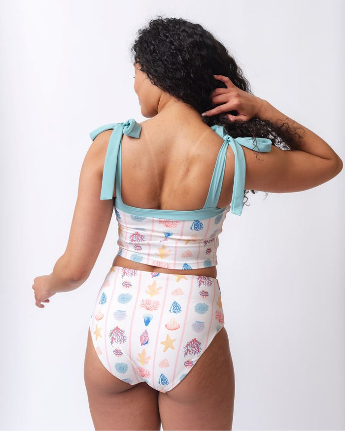 Photo of a woman wearing a seashell striped shoulder-tie swim crop top and a seashell striped high waist swim bottom- back angle