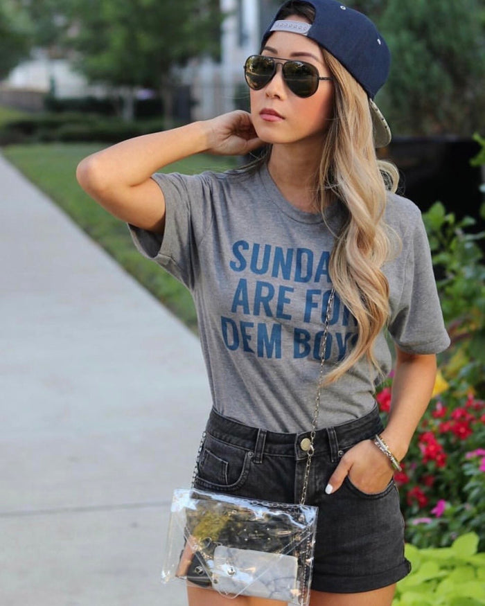 Woman wearing a Bullzerk "Sundays are for dem boys" tshirt, black jean shorts, and sunglasses with a backwards hat, standing outdoors.