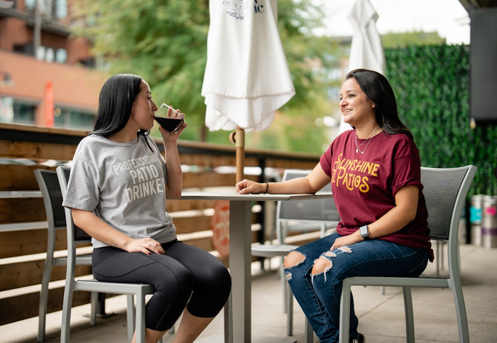 Two women sitting at an outdoor table, one drinking from a glass, the other engaged in conversation, both wearing Bullzerk tshirts.