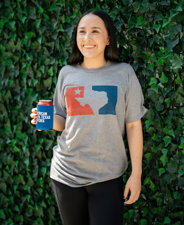 Woman standing in front of greenery, wearing a Bullzerk Texans Love Baseball tshirt and holding a canned drink in a koozie.