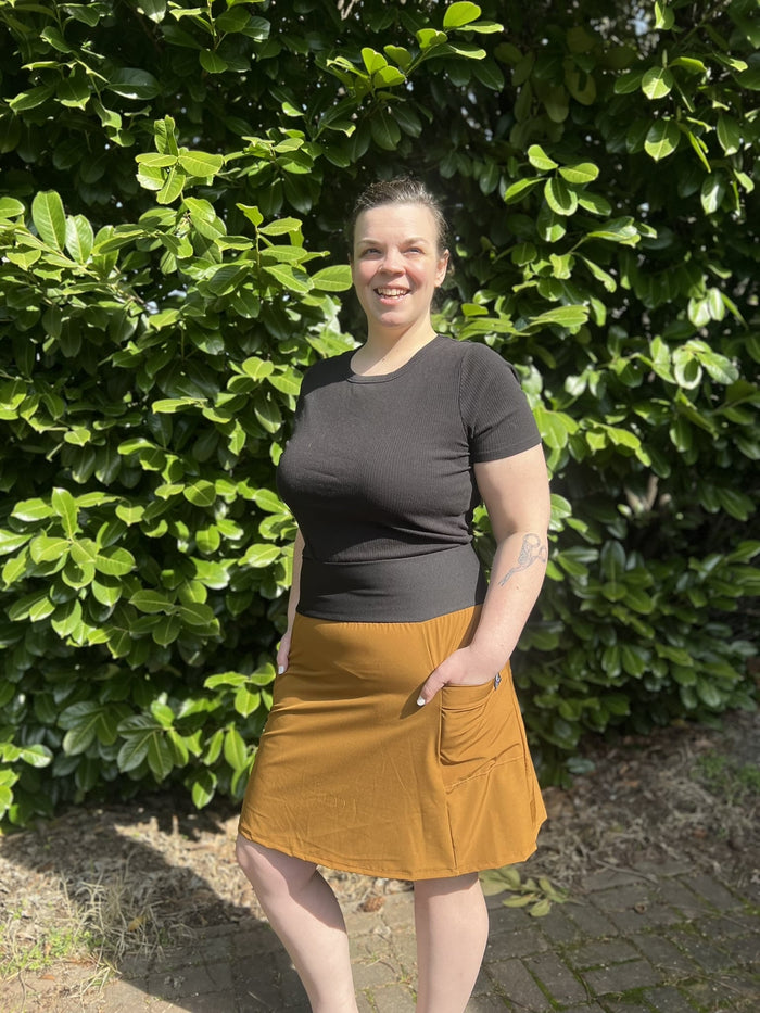 Person wearing a black top and copper skirt standing in front of green bushes.