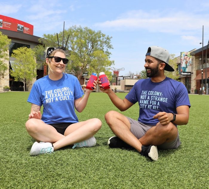 Two people laughing and sitting on a lawn wearing Bullzerk tshirts and clinking two drinks together.