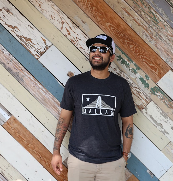 Man standing in front of a wooden wall, wearing a Bullzerk Trinity tshirt, sunglasses and a baseball hat.