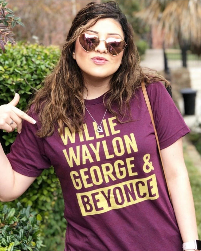 Woman wearing a Bullzerk "Willie Waylon George and Beyonce" tshirt and heart sunglasses, pointing to her shirt.