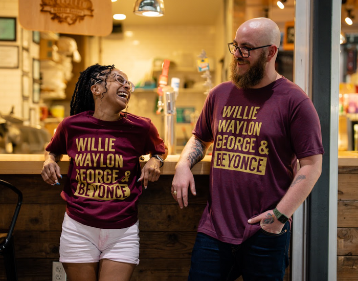 Two people wearing matching Bullzerk Willie Waylon George and Beyonce tshirts, leaning against a bar.